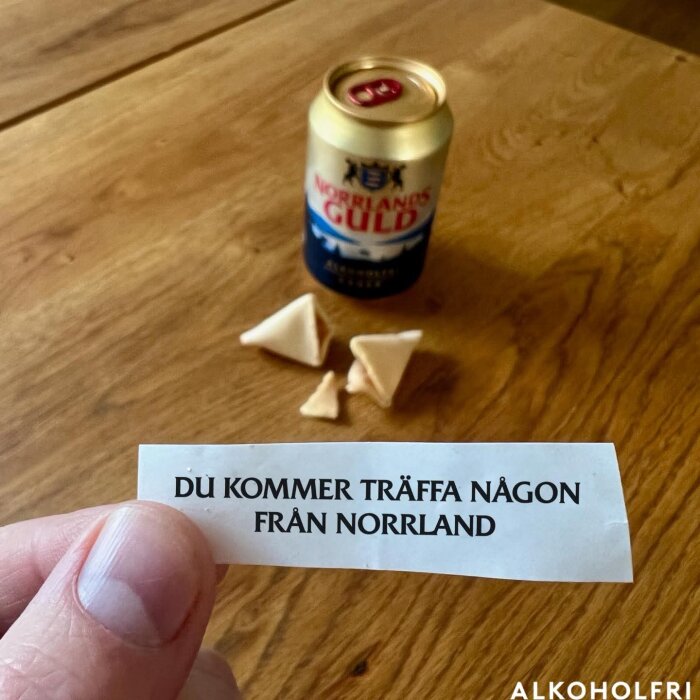 Lyckokaka med lapp som säger "DU KOMMER TRÄFFA NÅGON FRÅN NORRLAND" bredvid alkoholfri öl på träbord.