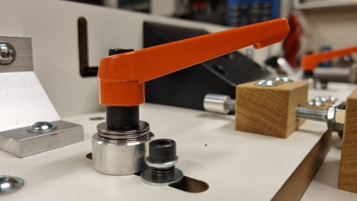 Närbild av en orange spännspak monterad på ett fräsbord mot en vit bakgrund med trä- och metallkomponenter.