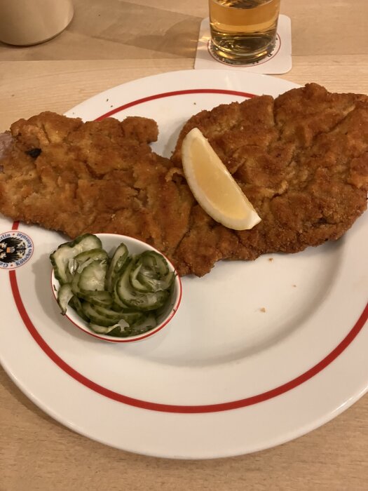 Wienerschnitzel med en citronskiva och skivad gurka på tallrik, med ett glas dryck i bakgrunden.
