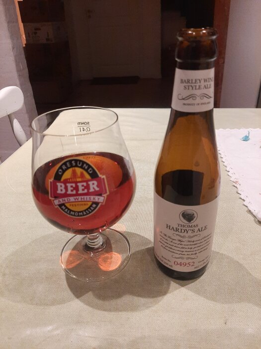 Glas med rödbrun öl bredvid flaska med etiketten "Thomas Hardy's Ale" på ett bord med vit duk.