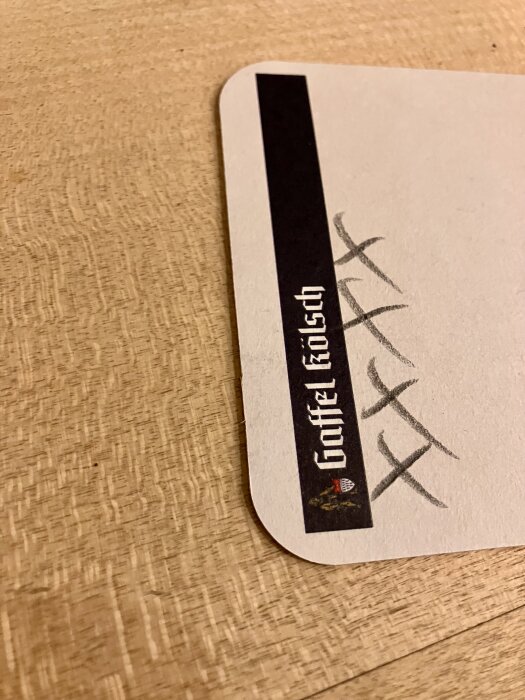 Ett ølunderlägg på ett träbord med texten "Gaffel Kölsch" och flera handritade kryss ovanpå underlägget.