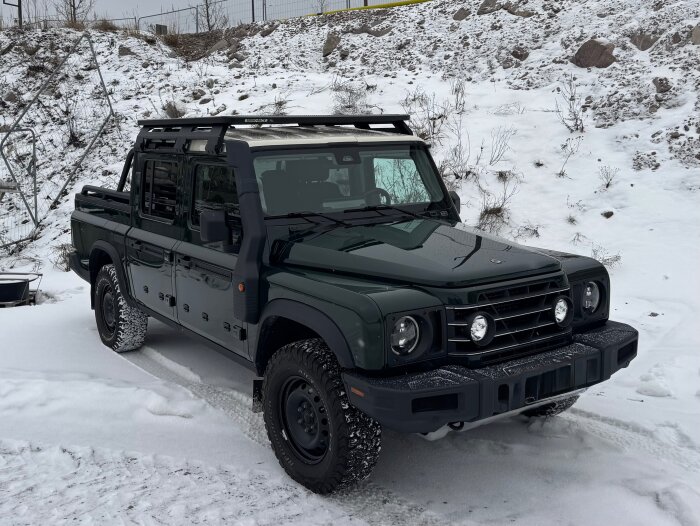 En mörkgrön off-road bil stående i ett snöigt landskap med snötäckta hjul och takräcke.