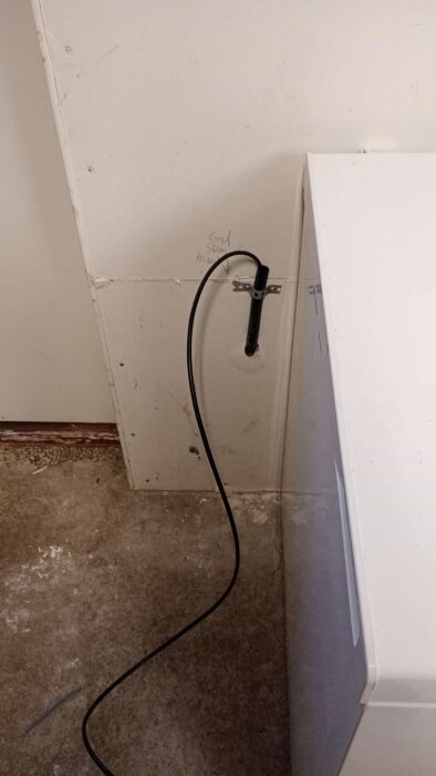 Svart kabel som kommer ut genom vit vägg, fäst med ett svart beslag. Grovt golv och skugga av vit möbel syns.