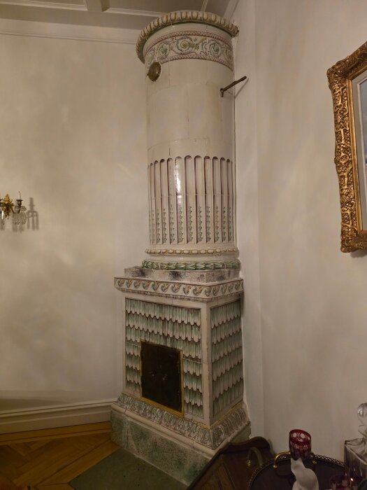 Dekorerad kakelugn i traditionell stil, placerad i ett hörn av ett rum med ljusa väggar och trädetaljer.