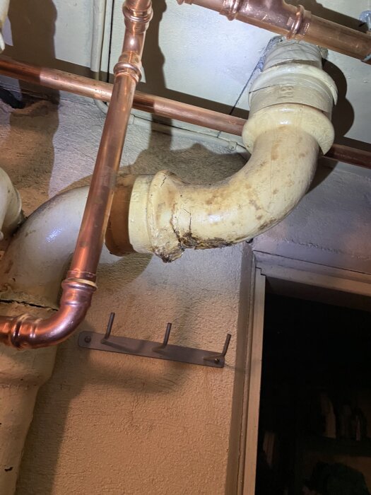 Rostigt avloppsrör av gjutjärn i källartak med kopparrör intill. Rörens skick och läckage orsak till oro vid renovering eller reparation.