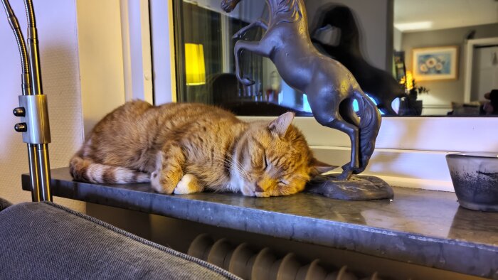 En orange katt sover på en fönsterbräda bredvid en skulptur av en häst.