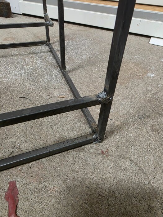 Ett svetsat vedställ av rostfria stänger. Fogarna ser ojämna ut, men det är hemgjort. Står på ett betonggolv framför en vit vägg.