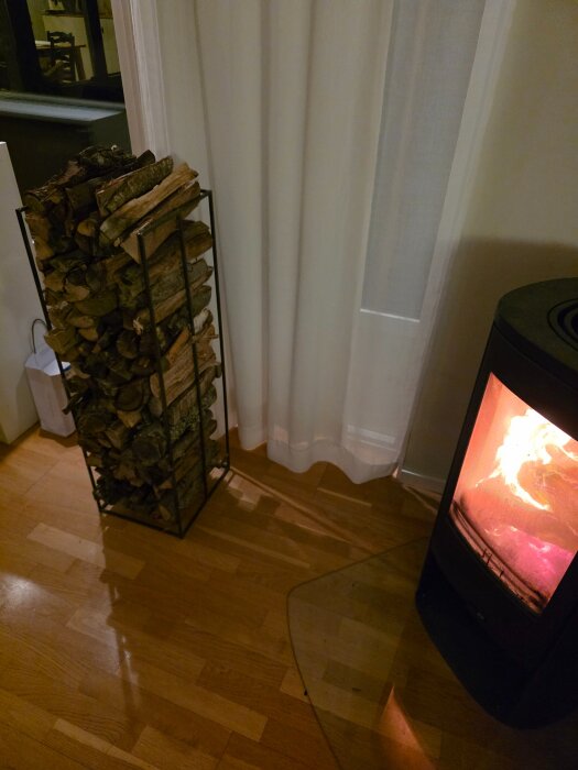 Rustikt vedställ fylld med ved bredvid en brinnande kamin, placerat på ett trägolv framför vita gardiner.
