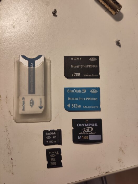 Olika typer av äldre minneskort på ett bord, inklusive Sony Memory Stick och Olympus xD-kort, omgivna av små skruvar.
