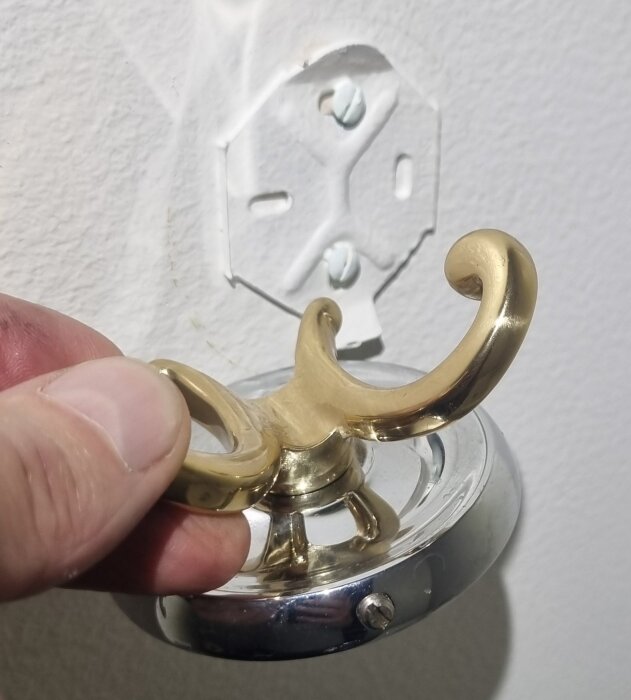 Hand som håller en mässingskrok med silverfäste och skruv i underkant, monterad på väggen.