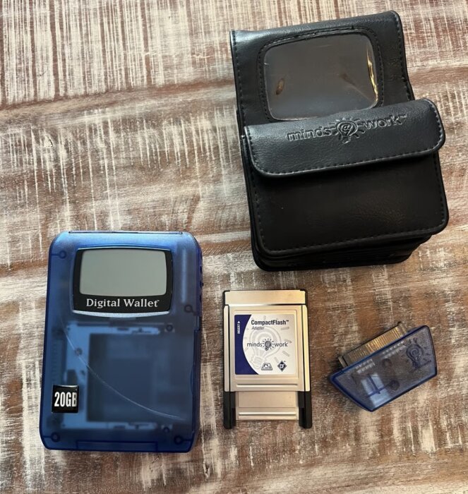Hårddiskbaserad bärbar lagringsstation "Digital Wallet" med tillbehör, inklusive CompactFlash Adapter och svart förvaringsväska, på träbord.