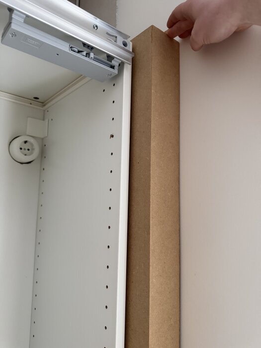 En hand placerar en trälist vid sidan av ett skåp, nära en dörrkarm, för att skapa ett grepp.