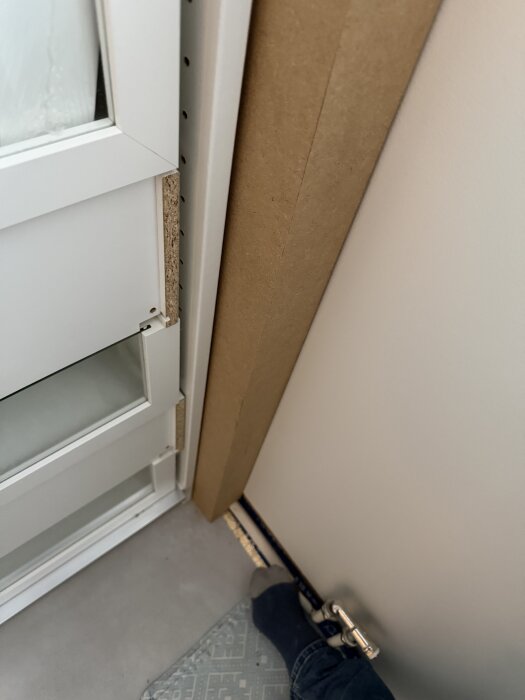 Bild på en dörrkarm med påbörjad montering av tröskel; detaljer av dörrens kant, tröskeln och en matta syns, med en fot i strumpa nära tröskeln.