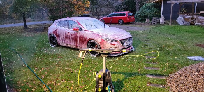 En röd bil täckt med tvålskum tvättas på en gräsmatta utan dagvattenbrunnar, intill en högtryckstvätt.