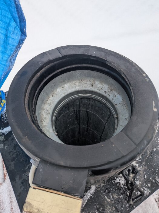 Överdel av en takfläkt med synlig fläns och insida av ventilationsrör, omgiven av snö och presenning.