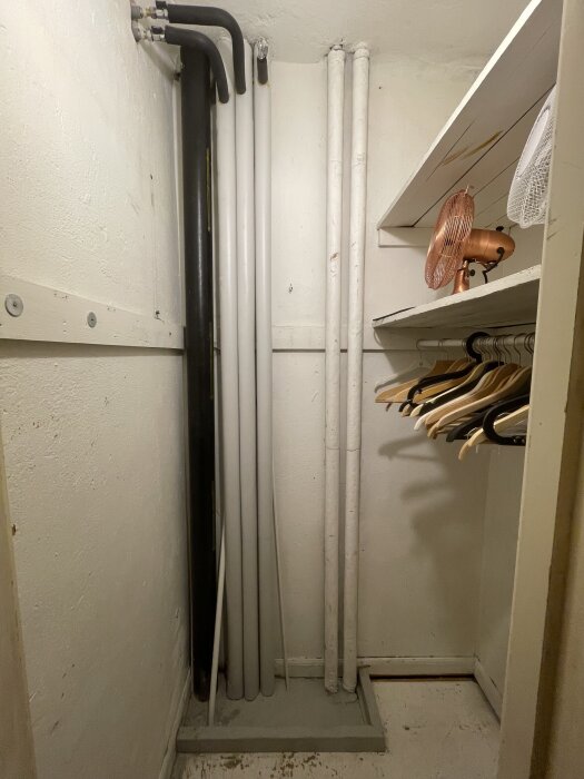 Rörinstallation i en klädkammare, med olika rör inklusive en svart avloppsstam och vita vattenrör, bredvid hyllor och galgar.