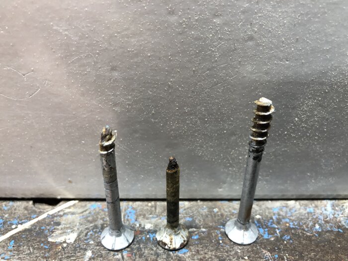 Tre skruvar placerade vertikalt på en träyta, vissa med rost. Bakgrunden är en putsad vägg.