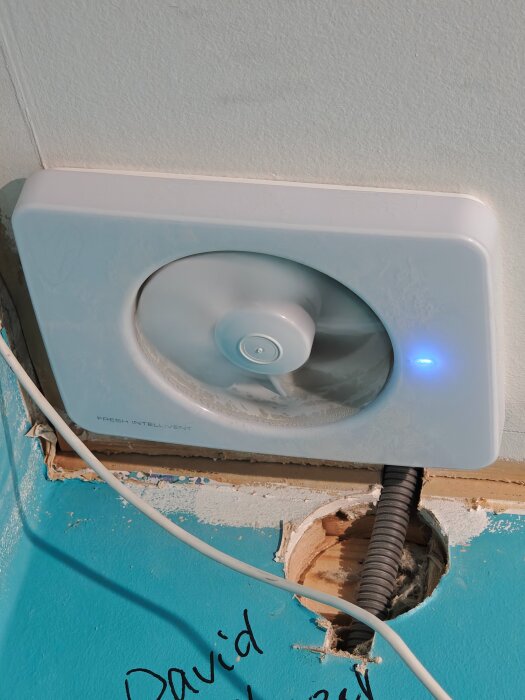Ventilationsfläkt på vägg med blått ljus, omgiven av smuts och en nedåtriktad sladd genom ett hål i golvet under installation.