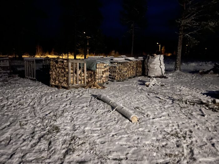Vedstaplar på snöigt fält nattetid, omgivna av träd i mörkret.