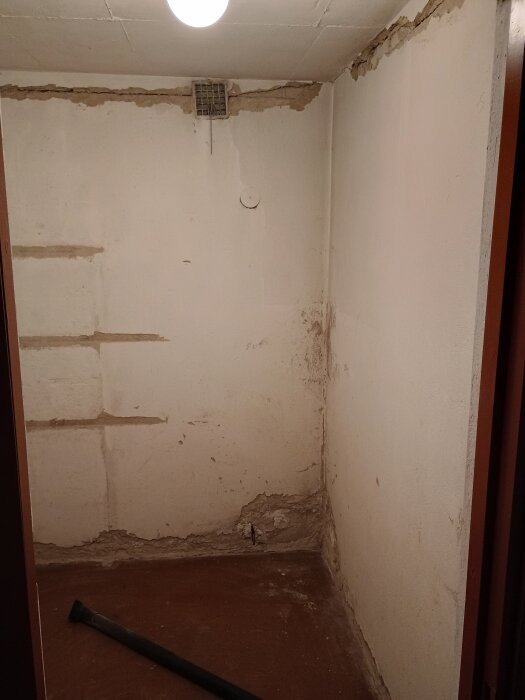 Nedgånget rum med avskalade väggar och blottlagt murbruk. En rörbit ligger på ett jordgolv. Plafondlampa i taket och ventilikationsöppning på väggen.