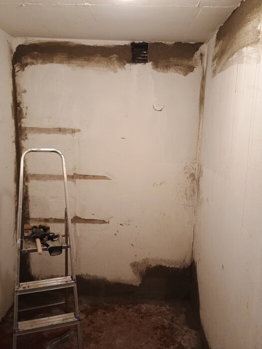 Vit inomhusvägg under renovering med fläckar av murbruk och en stege med verktyg. Ventilhål synligt på överkanten av väggen.