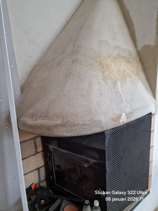 Öppen spis med vit och delvis skadad kåpa i ett kök, synlig murstock med sprickor och metallnät framför, samt diverse föremål på spishyllan.