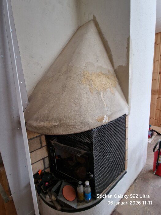 Gammal spis med konisk kåpa och brandskador ovanför eldstad, omgiven av väggar med sprickor; renoveringsmaterial syns på spisen.