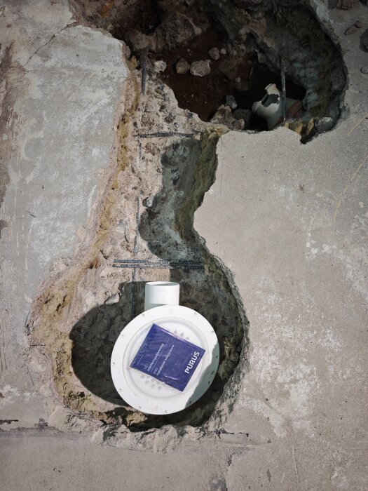 Hål i golvet med en installerad golvbrunn och en manual från Purus, omgiven av betongbeklädd yta.