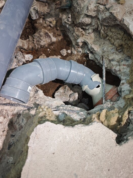 Sprucket rör i hål i betonggolv, omgivet av grus och isolering. Röret är skadat och behöver åtgärdas under byggprojekt.