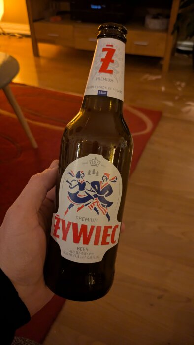 En hand håller i en flaska polskt öl, Żywiec, med en etikett som visar en dansande duo. Inredningen i bakgrunden är varmt belyst.