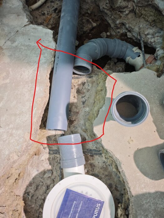 Avloppsrör i betonggolv med synliga ledningar och en vit dräneringsenhet. En röd penna markerar området där rören ska anslutas.