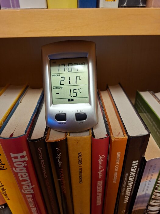 Digital termometer visar inomhustemperatur 21,1°C och utomhustemperatur -1,5°C, placerad på en bokhylla med flerfärgade böcker.