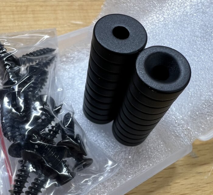Svarta magneter staplade i två rader bredvid plastförpackning med skruvar på ett bord.