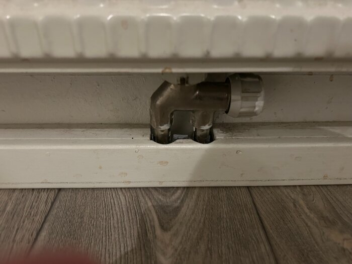 Luftningsnippel på radiator som bör bytas ut, monterad mot en vägg över trägolv.