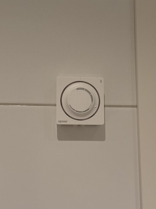 Väggmonterad Uponor termostat med en inställningsratt i neutralt läge på vit kakelvägg.