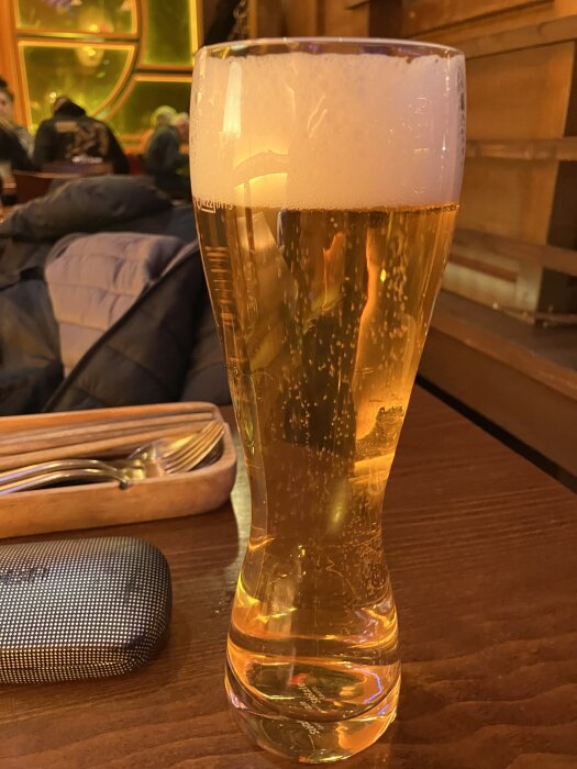 Ett glas öl med skum på toppen står på ett träbord i en varm atmosfär med andra besökare och bestick i bakgrunden.