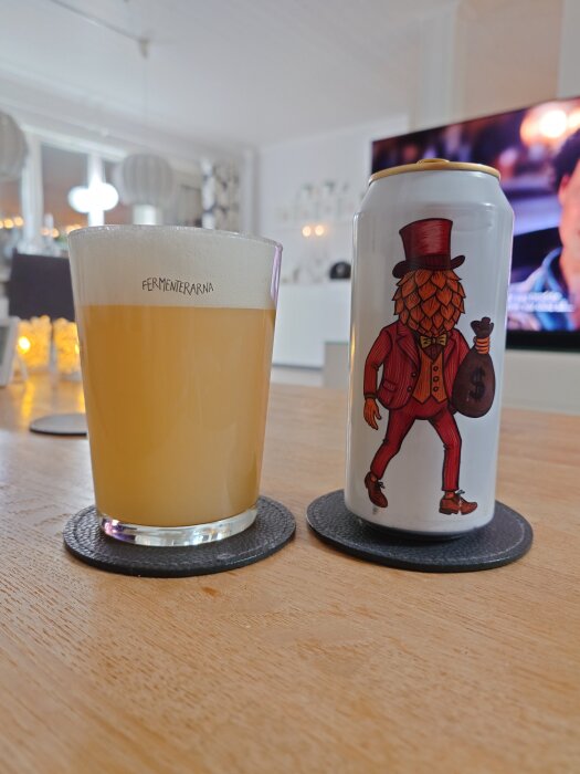 Glas med ljus öl från Fermenterarna bredvid en burk med illustration av en figur med kottansikte och hatt på ett bord inomhus.