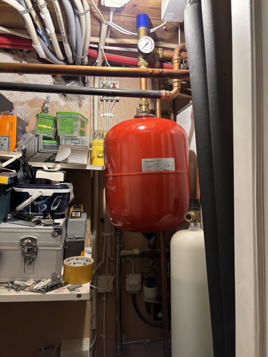 Rött expansionskärl installerat i ett förråd, omgivet av rör och olika byggverktyg på hyllor.