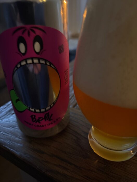 Bröl West Coast IPA med färgglad etikett och ett glas delvis fyllt med skummande öl på ett träbord.