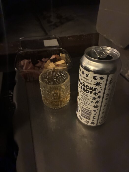 En burk öl, ett glas med dryck och en plastförpackning med chark och ostbitar på ett bord i svag belysning.