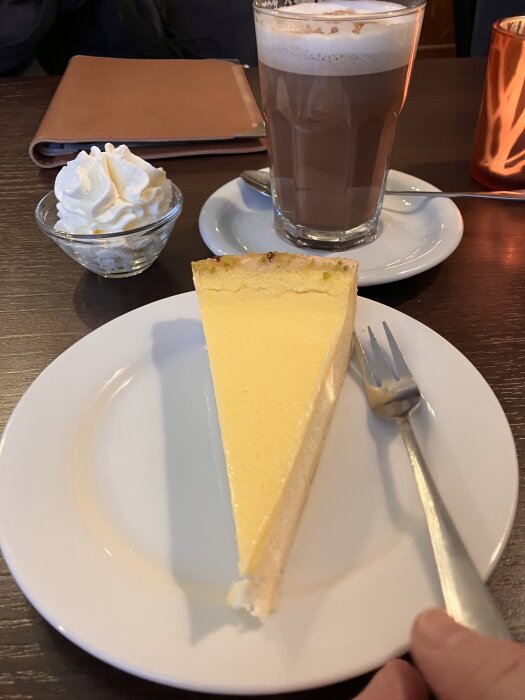 En bit ostkaka på en vit tallrik, med vispad grädde och varm choklad i en glas bredvid.