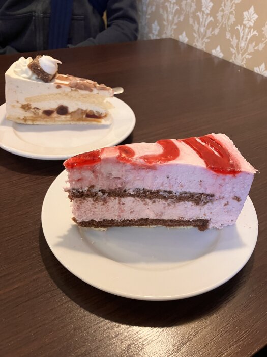 Två tårtbitar på vita tallrikar på ett bord, en med rosa grädde och jordgubbssås, en med vit grädde och choklad.
