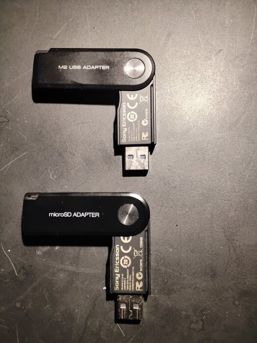 Två Sony Ericsson USB-adaptrar på ett bord, en för M2 och en för microSD, visas för att illustrera anslutning med standard USB.