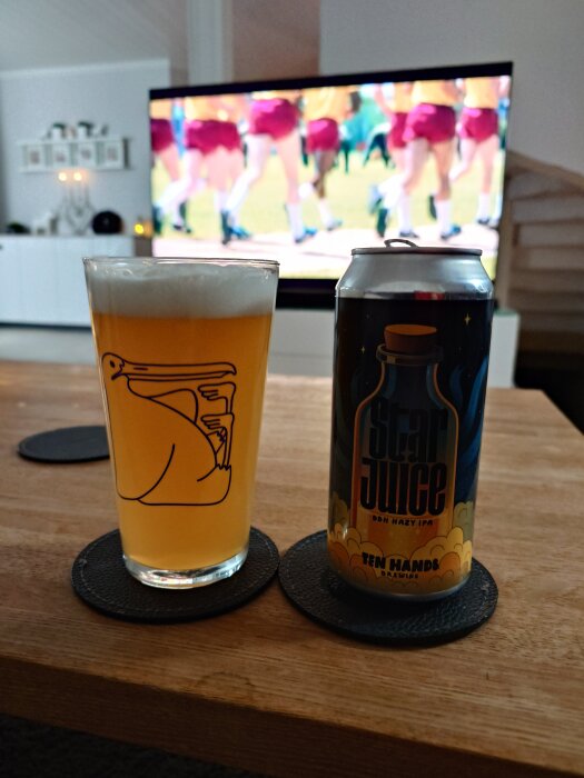 En ölburk med texten "Star Juice" och "Ten Hands Brewing" står bredvid ett glas öl på ett bord, med en TV i bakgrunden.
