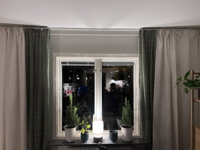 Fönster med två gardinpar, krukväxter på fönsterbrädan och en lampa i mitten, tänd i ett mörkt rum.