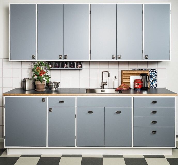 Modulkök med blå skåpluckor, vit kakelvägg, och synliga skarvar. Svart bänkskiva, diskho, och detaljer som prydnadsväxter och köksutrustning.