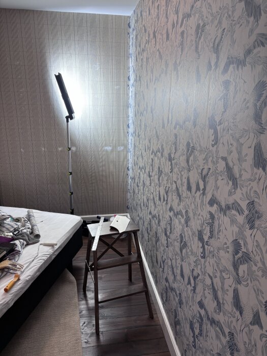 Bygglampa lyser mot vägg med blommig tapet, inställd på 50% effektläge. Verktyg och tapetmaterial på en stegpall och säng i rummet.
