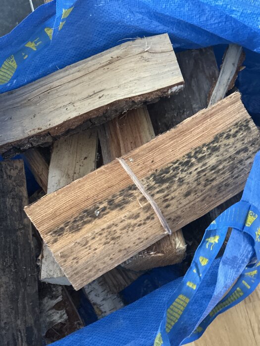 Bitar av blandved med mörka fläckar liggande i en blå säck, ifrågasätts om de är lämpliga att elda i kakelugn.