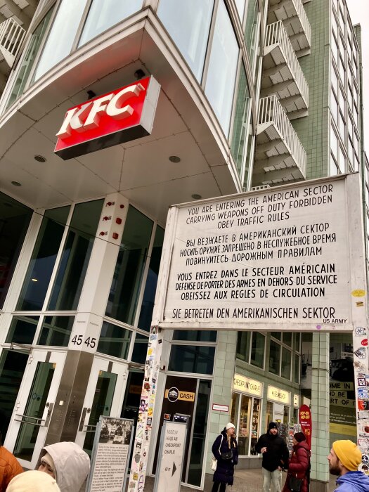 Folk vid en byggnad med ett KFC-skylt, samt en skylt med text om att man går in i den amerikanska sektorn, vid Checkpoint Charlie i Berlin.