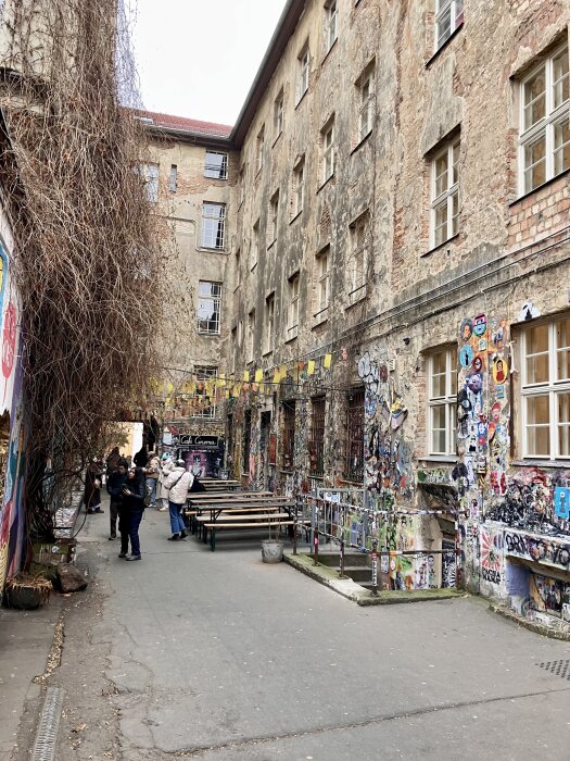 Trång gränd med gamla byggnader täckta av klistermärken och graffiti, träbänkar och några människor som går förbi eller står still.